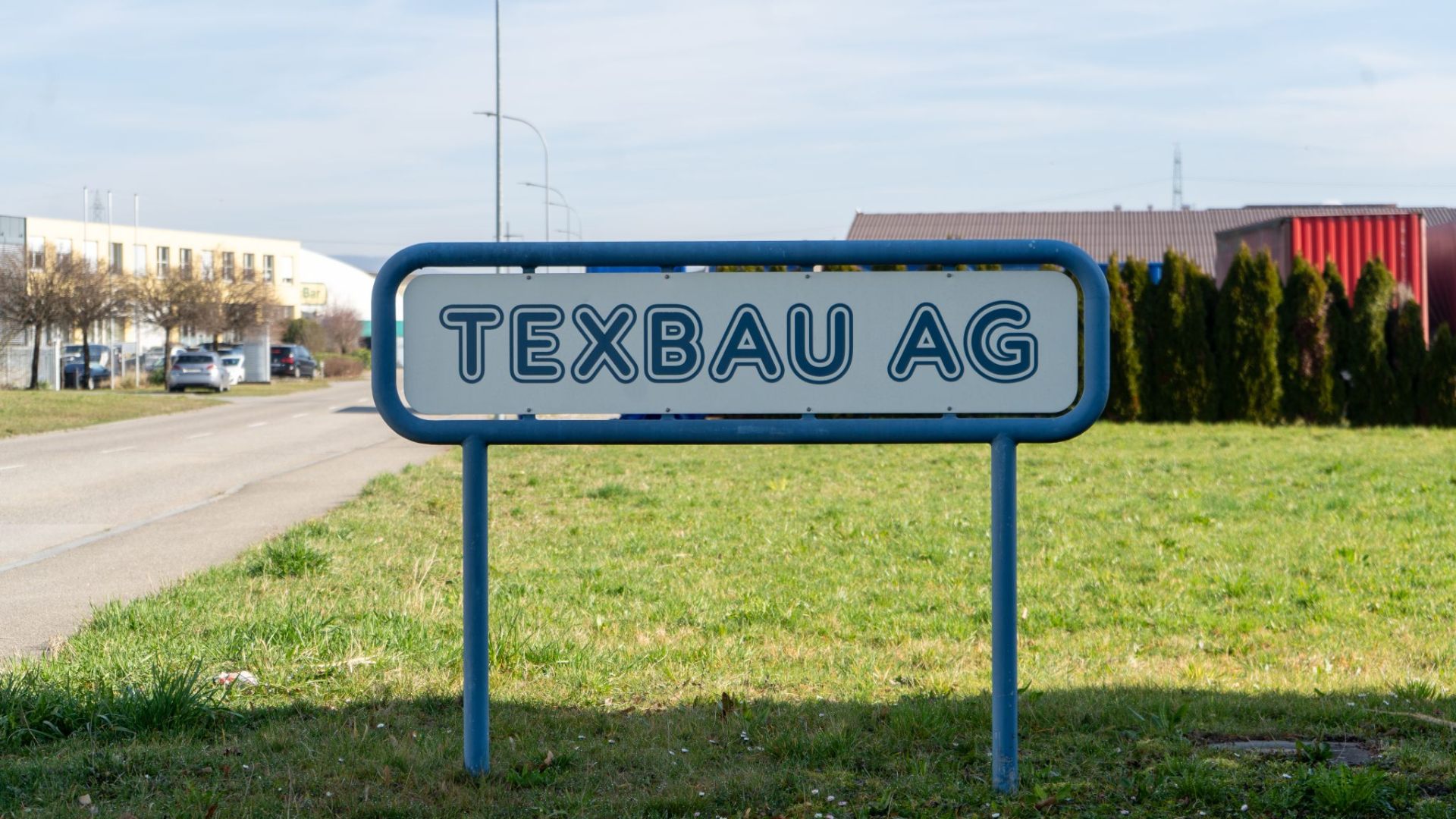 Blau umrahmtes Firmenschild „Texbau AG“ am Strassenrand in Däniken.