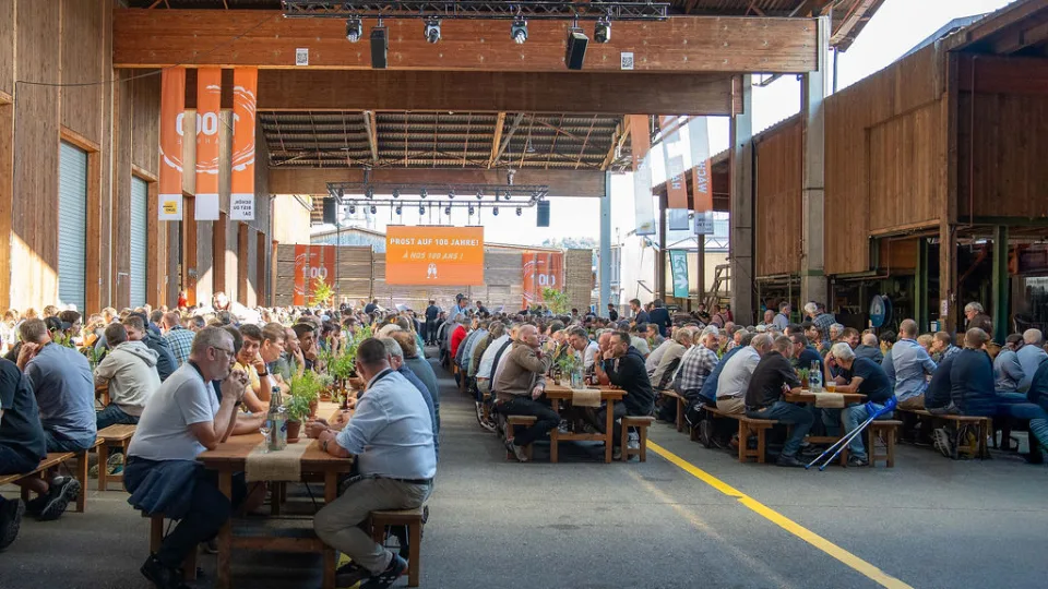 Lange Reihen von Festbänken in einer offenen Holzhalle, voll besetzt mit Gästen, im Hintergrund ein oranges Banner mit der Aufschrift «Prost auf 100 Jahre!».
