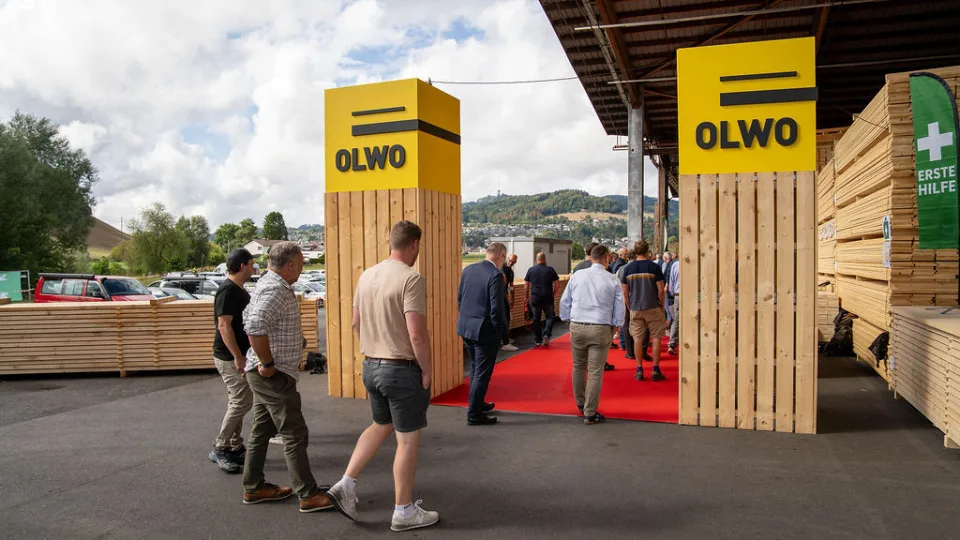 Roter Teppich zwischen zwei Holzpfeilern mit gelbem OLWO-Logo, Gäste gehen in die Halle, rechts gestapeltes Holz.