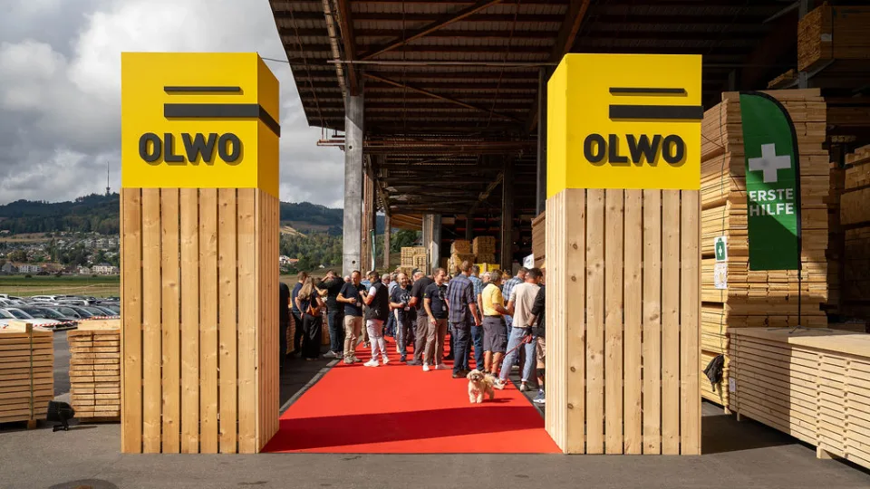 Blick über den roten Teppich in die Halle: Gäste zwischen gestapeltem Holz, im Vordergrund ein Pfeiler mit OLWO-Logo und ein kleiner Hund.
